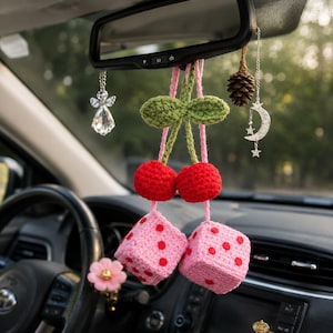 May include: A car interior with various hanging decorations. A pair of red crocheted cherries with green leaves and pink stems hangs from the rearview mirror, along with pink dice with red dots. Additional charms include a crystal angel, a pinecone, and a silver moon and stars.
