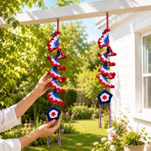 May include: Two crochet wind chimes with red, white, and blue spiral designs and star-shaped bottoms. The wind chimes are hanging outdoors, with a white pergola and green foliage in the background. Each star has a white center and red border.