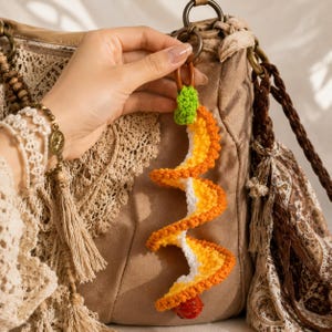May include: A handmade crochet keychain in a spiral design, featuring orange, yellow, and white yarn. The keychain has a small green leaf and a red bell at the bottom. The keychain is attached to a brown leather loop.