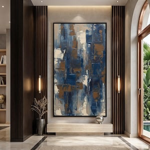 May include: Large abstract painting with a black frame. The artwork features a textured design with shades of blue, white, and brown. The painting is displayed in a modern interior setting with a neutral color palette and warm lighting.