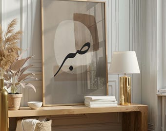 May include: Framed abstract art print with a gold frame. The artwork features a beige and brown color palette with a black Arabic calligraphy design. The print is displayed on a wooden bench with a gold lamp and a plant.