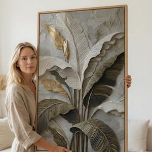 May include: A rectangular wall art piece featuring a textured, three-dimensional relief of large tropical leaves in shades of sage green, cream, and metallic gold, all encased within a light-toned wooden frame with a visible grain pattern.