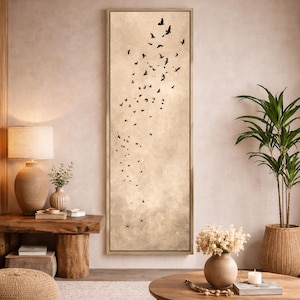 May include: A framed vertical canvas art piece featuring a flock of black birds in flight against a beige, cloudy background. The artwork is in a gold frame and is displayed in a room with neutral tones and natural elements.