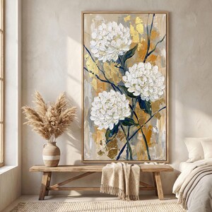 May include: A framed floral painting featuring three large white flowers with textured petals. The artwork has a neutral color palette with gold leaf accents and dark blue stems against a beige and gold background.