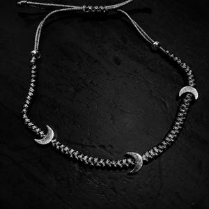May include: Black and white necklace featuring three silver crescent moon charms. The necklace is made of a woven black and white cord with adjustable length. The background is a dark, textured surface.