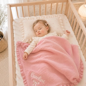 May include: A sleeping baby in a wooden crib, covered with a pink ruffled blanket. The blanket has the name "Caroline" embroidered in white. The crib has a floral patterned sheet and pillow.