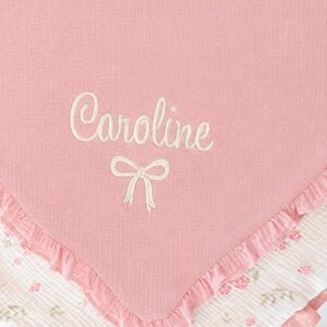 May include: A pink baby blanket with the name "Caroline" embroidered in white script, along with a bow. The blanket has a ruffled edge and a floral patterned border in pink and white.