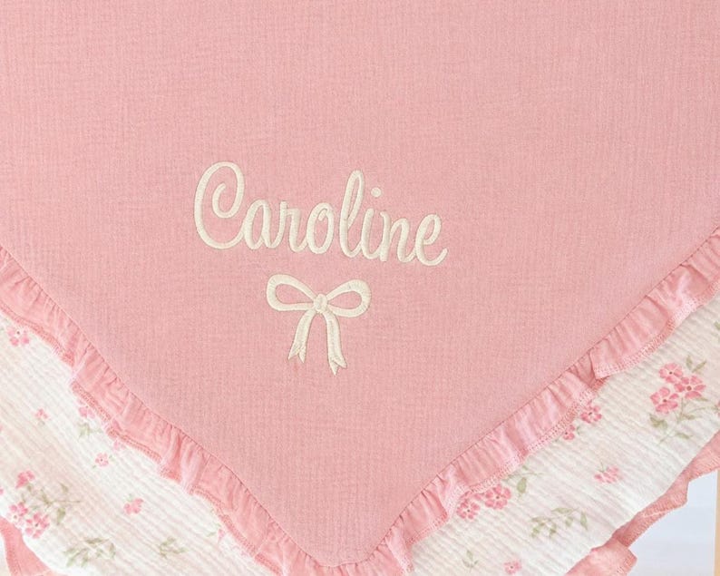 May include: A pink baby blanket with the name "Caroline" embroidered in white script, along with a bow. The blanket has a ruffled edge and a floral patterned border in pink and white.