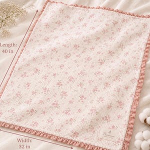 May include: A rectangular, floral-patterned blanket with a pink ruffled edge. The blanket is 40 inches long and 32 inches wide. The pattern features small pink flowers on a cream background. A small label in the corner reads "Premium".