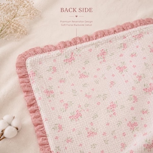May include: A reversible baby blanket with a floral pattern. The blanket features a soft, white fabric with small pink flowers and green leaves. The border is a ruffled, dusty rose color. The text "BACK SIDE" is visible.