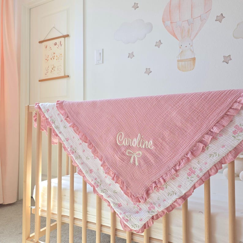 May include: A pink baby blanket with the name "Caroline" embroidered in white, draped over a wooden crib. The blanket has a ruffled edge and a floral patterned border. A nursery wall features a hot air balloon and star decals.