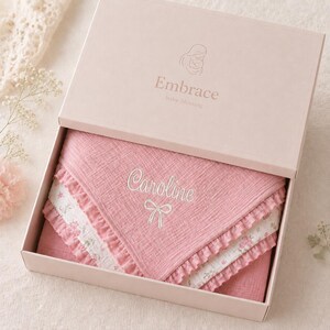 May include: A pink baby blanket with the name "Caroline" embroidered in white, presented in a matching box. The blanket features a ruffled edge and floral patterned trim. The box lid has the word "Embrace" and a baby graphic.