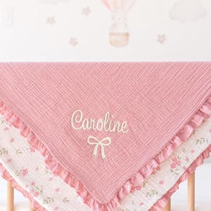 May include: A pink baby blanket with the name "Caroline" embroidered in cursive script, accented by a bow. The blanket features a ruffled edge and a floral patterned border. The blanket is draped over a crib.