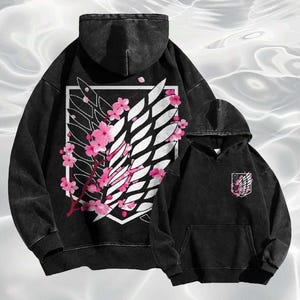 May include: Two black hoodies with a faded wash effect. The larger hoodie features a white emblem with a wing design and pink cherry blossoms. The smaller hoodie has a pocket with a matching emblem. Both have hoods.