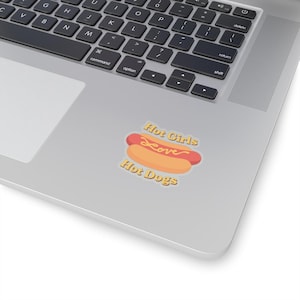 Puede incluir: Una pegatina con un perrito caliente de dibujos animados con una salchicha roja y un panecillo amarillo. El texto "Hot Girls Love Hot Dogs" está escrito en letras cursivas y de bloque amarillas.