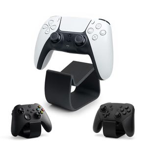 May include: A white and black gaming controller sits on a black stand, with two additional black controllers below. The controllers have various buttons and joysticks. The stand is curved and designed to hold the controller.