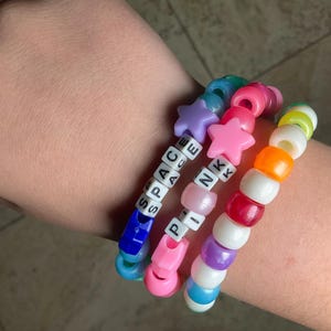 May include: Three colorful beaded bracelets. One bracelet spells "SPACE" with white letter beads and has blue and purple star beads. The second bracelet spells "PINK" with pink star beads. The third bracelet has a mix of colorful beads.