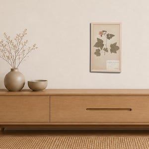 May include: A light wood media console with a long drawer and a recessed handle. On top sits a round beige vase with thin branches and a small beige bowl. A framed botanical print of a plant with green leaves and a pink flower hangs above.