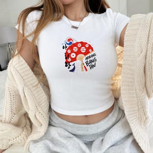 May include: White crop top with a graphic of a red fan with white circles and the text "Arirang Bang Tan!" in black. The fan design includes Korean script and a blue and red emblem. The shirt has short sleeves.