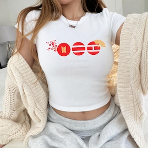 May include: White crop top with a graphic design featuring red circles and a fan. The design includes a floral element and a logo. The top is short-sleeved.