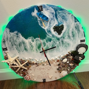 Puede incluir: Reloj de pared redondo con diseño de paisaje marino. La esfera del reloj presenta un océano azul con olas blancas, un pequeño bote azul y una isla en forma de corazón. El reloj está decorado con conchas marinas, estrellas de mar y los nombres "Pamela" y "Erblig".