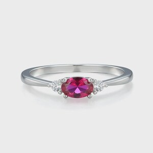 Op de afbeelding: Een zilveren ring met een grote, ovaal geslepen, magenta gekleurde edelsteen, geflankeerd door twee kleinere, ronde, heldere edelstenen. De ring heeft een eenvoudige, elegante band, perfect voor dagelijks gebruik of speciale gelegenheden.
