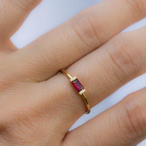 Puede incluir: Un anillo de oro con una piedra preciosa rectangular de color rojo intenso. El anillo tiene pequeñas piedras de acento transparentes a cada lado del rubí. El anillo está en un dedo, mostrando el diseño y el color de la joya.