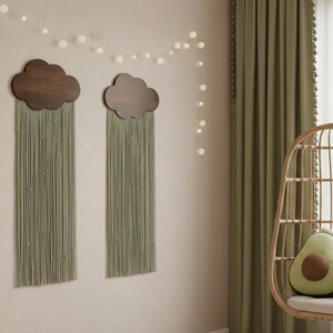 May include: Two wooden cloud wall hangings with olive green fringe are mounted on a beige wall. A string of white lights hangs above them. A wicker hanging chair with avocado-shaped pillows sits near a green curtain.