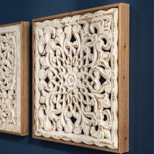 Könnte beinhalten: Ein quadratisches Wandpaneel aus weiß geschnitztem Holz mit einer abgenutzten Oberfläche, die darunter liegende natürliche Holztöne freilegt. Das Paneel ist in einen einfachen, hellen Holzrahmen gefasst und zeigt ein symmetrisches, florales Muster.