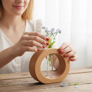 May include: A wooden ring-shaped vase holding a small bouquet of blue flowers. The vase has a clear glass tube for the flowers and is set on a wooden table. The vase is being held by a person with red nail polish.