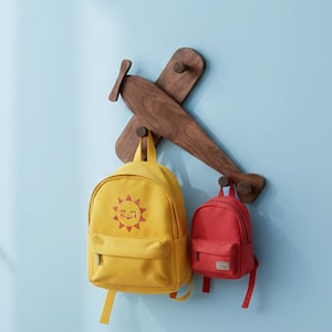 Op de afbeelding: Een houten vliegtuigvormige wandhaak met twee rugzakken eraan. De haak is gemaakt van donker hout en is bevestigd aan een lichtblauwe muur. Een gele rugzak met een lachende zon en een rode rugzak hangen aan de haken.