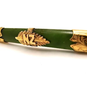 May include: A cylindrical green stone bar brooch featuring gold-toned metal end caps with engraved leaf patterns and a central gold-toned metal emblem shaped like a fern leaf with the letters NZ embossed in the centre of the leaf design.