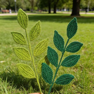 May include: Two crochet leaf sprigs in shades of green. One is a lighter, yellowish-green, and the other is a darker, forest green. The leaves have detailed stitching, and the stems are thin and wire-like. The background is a blurred green lawn.