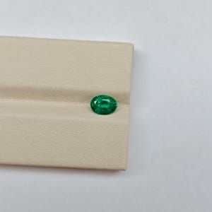 Pode incluir: Uma gema verde de corte oval com uma superfície facetada que reflete a luz. A pedra exibe um tom esmeralda profundo e saturado e está posicionada num suporte de exibição bege claro texturizado com um sulco horizontal no centro.