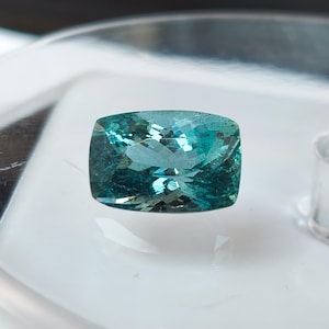 Può includere: Una pietra preziosa taglio cuscino con un vibrante colore blu-verde e una superficie sfaccettata che riflette la luce. La pietra ha una forma rettangolare con angoli arrotondati e mostra una chiarezza interna con rifrazione della luce.