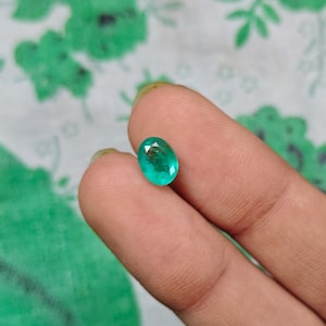 Può includere: Una piccola gemma sfaccettata di forma ovale con un colore verde vibrante e traslucido. La pietra presenta una superficie lucida con inclusioni interne visibili e un taglio brillante che riflette la luce sui suoi bordi arrotondati.