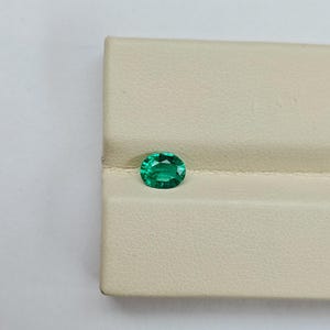 Pode incluir: Uma gema verde de corte oval com uma superfície facetada que reflete a luz. A pedra apresenta um tom esmeralda profundo e saturado, posicionada sobre uma superfície de exibição bege clara e texturizada com um vinco horizontal.