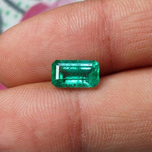 Puede incluir: Una piedra preciosa esmeralda rectangular con un tono verde vibrante y una superficie facetada. La piedra presenta un acabado pulido y claro con inclusiones internas visibles, mostrando un diseño clásico de talla esmeralda.