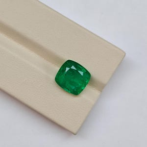 May include: A cushion-cut emerald gemstone with a vibrant green hue and faceted surface. The gemstone displays internal clarity characteristics and light reflection across its polished top, showcasing a square shape with rounded corners.