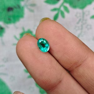 Pode incluir: Uma pedra preciosa de esmeralda de corte oval com um tom verde vibrante e translúcido e uma superfície multifacetada. A pedra apresenta clareza interna e facetas que refletem a luz, exibindo uma cor verde brilhante e saturada.