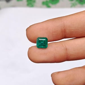 May include: A square-cut emerald gemstone with a deep green hue and faceted surface. The stone displays a clear, transparent quality with visible internal inclusions and sharp, defined edges, showcasing a classic geometric shape.