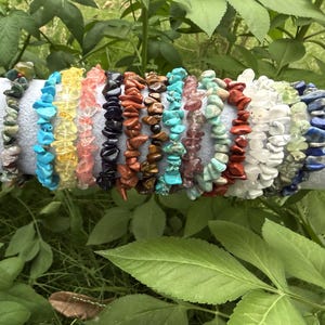 May include: A collection of gemstone chip bracelets in various colors, including turquoise, red, and white. The bracelets are strung on elastic and displayed on a white cylindrical surface, surrounded by green foliage.