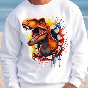 Könnte beinhalten: Weißes Sweatshirt mit einem lebendigen orangefarbenen Tyrannosaurus Rex, der durch eine Wand bricht. Die Grafik des Dinosauriers ist von bunten Farbspritzern in Rot, Gelb und Blau umgeben. Ein lustiges und auffälliges Design.