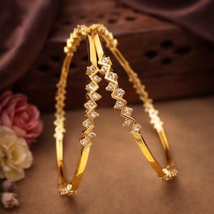 Op de afbeelding: Twee gouden armbanden met een zigzagpatroon van vierkante, heldere edelstenen. De armbanden worden gepresenteerd op een bruin oppervlak met een vage achtergrond van een houten kist en bloemen. De armbanden hebben een glanzende, gepolijste afwerking.