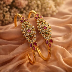 May include: Two gold bangles with intricate designs. Each bangle features a central square with a red gemstone, surrounded by clear stones and smaller green and red gems. The bangles rest on a peach-colored fabric.