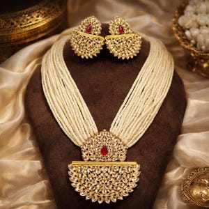 May include: A gold-toned necklace and earring set with a cream-colored beaded strand. The necklace features a large pendant with a red gemstone center. The earrings have a similar design with a red gemstone. The jewelry is displayed on a brown stand.