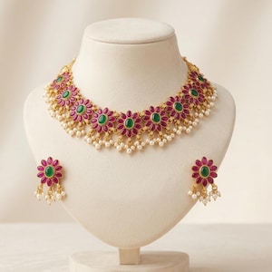 Könnte beinhalten: Ein goldfarbenes Halsketten- und Ohrring-Set. Die Halskette und die Ohrringe haben ein Blumenmuster mit rubinroten Blütenblättern, grünen Zentren und baumelnden weißen Perlen. Der Schmuck wird auf einer weißen Schaufensterpuppe präsentiert.
