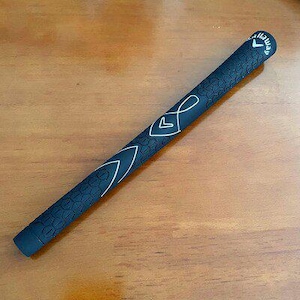 May include: A black golf club grip with white accent lines and the Callaway logo. The grip has a textured pattern and is lying on a wooden surface. The grip is approximately 10 inches long.