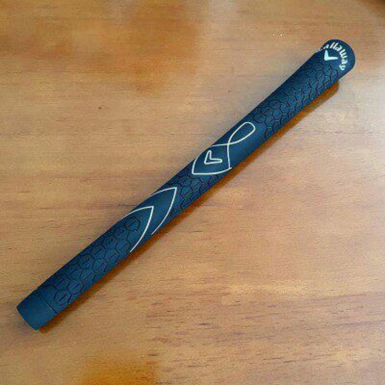 May include: A black golf club grip with white accent lines and the Callaway logo. The grip has a textured pattern and is lying on a wooden surface. The grip is approximately 10 inches long.