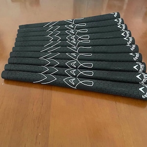 May include: A collection of black golf club grips with white accent designs. The grips have a textured surface and the brand name "Callaway" is visible on the end of each grip. The grips are arranged in a neat, organized stack.